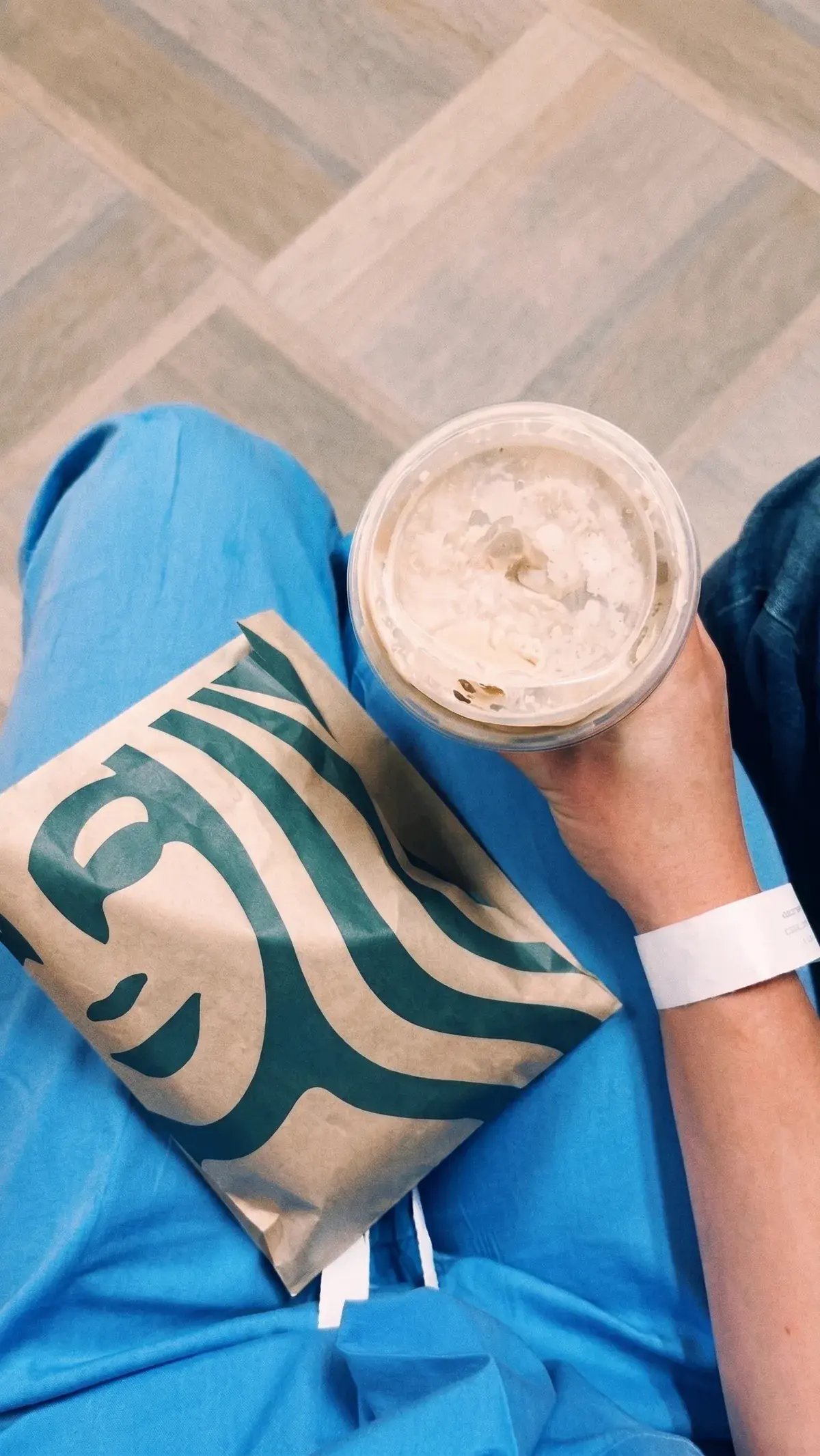 Overhead view of a healthcare worker in blue scrubs and a hospital wristband holding an iced Frappuccino with a paper food bag resting on their lap.