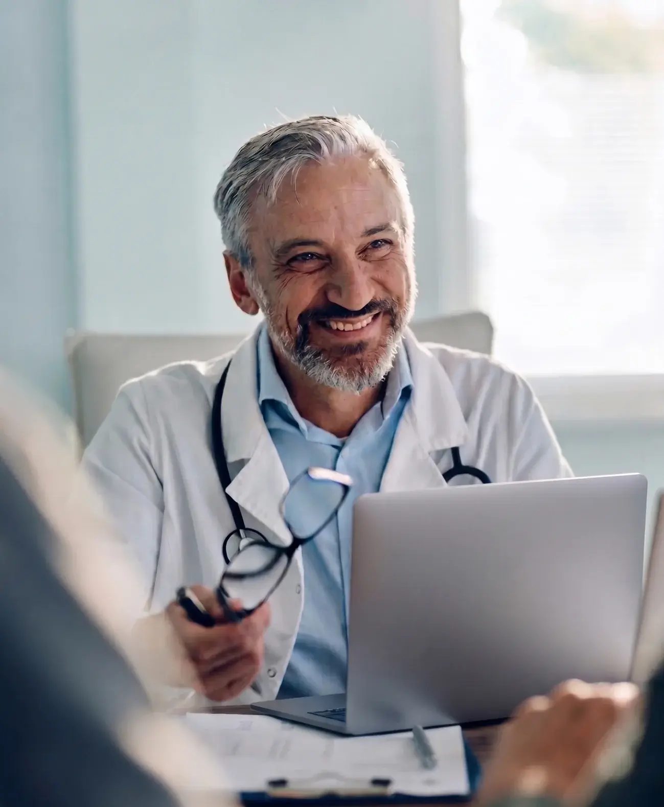 Physician in a white coat with a laptop, talking with a patient across the desk